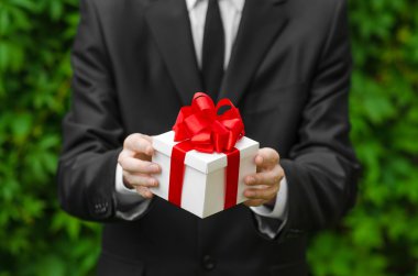 Hediye ve iş Tema: yeşil çim bir arka plan üzerinde kırmızı kurdele ile beyaz bir kutu içinde bir hediye tutarak siyah takım elbiseli bir adam