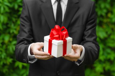 Hediye ve iş Tema: yeşil çim bir arka plan üzerinde kırmızı kurdele ile beyaz bir kutu içinde bir hediye tutarak siyah takım elbiseli bir adam