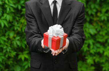 Gift and business theme: a man in a black suit holding a gift in a red box with a white ribbon on a background of green grass
