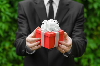 Gift and business theme: a man in a black suit holding a gift in a red box with a white ribbon on a background of green grass