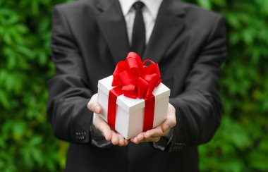 Hediye ve iş Tema: yeşil çim bir arka plan üzerinde kırmızı kurdele ile beyaz bir kutu içinde bir hediye tutarak siyah takım elbiseli bir adam