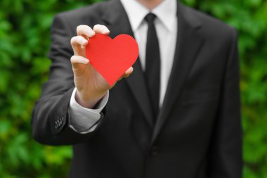 Love and business theme: a man in a black suit holding a card in the shape of a red heart on the background of green grass
