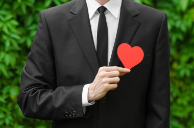 Love and business theme: a man in a black suit holding a card in the shape of a red heart on the background of green grass