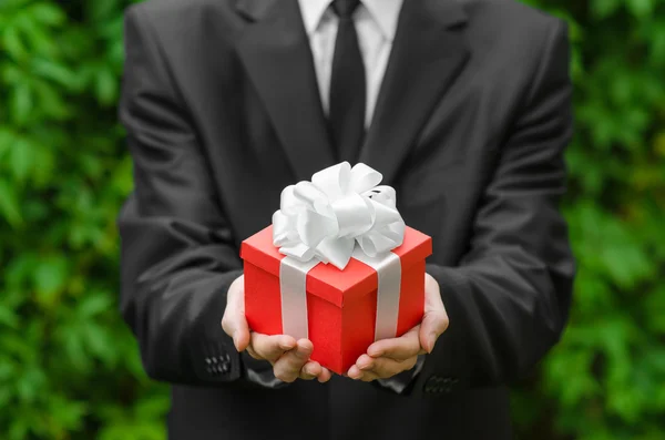 Tema hadiah dan bisnis: seorang pria dalam setelan hitam memegang hadiah di kotak merah dengan pita putih di latar belakang rumput hijau — Stok Foto