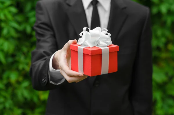 Gift and business theme: a man in a black suit holding a gift in a red box with a white ribbon on a background of green grass — Zdjęcie stockowe