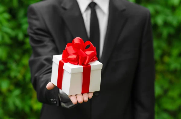 Regalo y tema de negocios: un hombre con un traje negro sosteniendo un regalo en una caja blanca con una cinta roja sobre un fondo de hierba verde —  Fotos de Stock