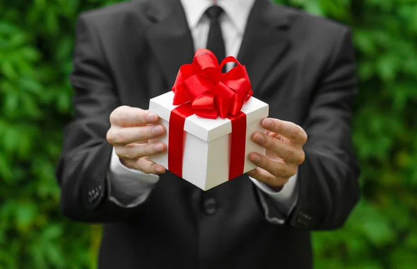 Tema hadiah dan bisnis: seorang pria dalam setelan hitam memegang hadiah dalam kotak putih dengan pita merah di latar belakang rumput hijau — Stok Foto