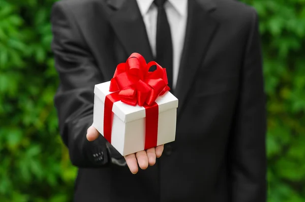 Gift and business theme: a man in a black suit holding a gift in a white box with a red ribbon on a background of green grass