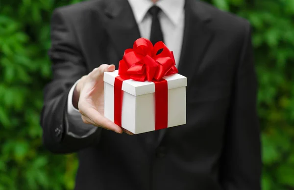 Regalo y tema de negocios: un hombre con un traje negro sosteniendo un regalo en una caja blanca con una cinta roja sobre un fondo de hierba verde —  Fotos de Stock