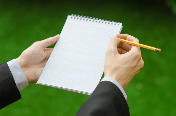 Day of knowledge and business topic: the hand of man in a black suit holding a notebook and pencil top view on a background of green grass — 스톡 사진