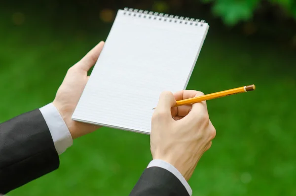 Day of knowledge and business topic: the hand of man in a black suit holding a notebook and pencil top view on a background of green grass — 스톡 사진