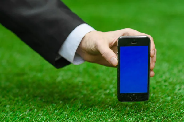 Comunicación y Negocios Asunto: Mano en un traje negro sosteniendo un teléfono moderno con pantalla azul en el fondo de hierba verde — Foto de Stock