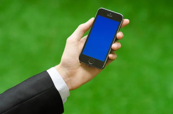 Communication and Business Subject: Hand in a black suit holding a modern phone with blue screen in the background of green grass