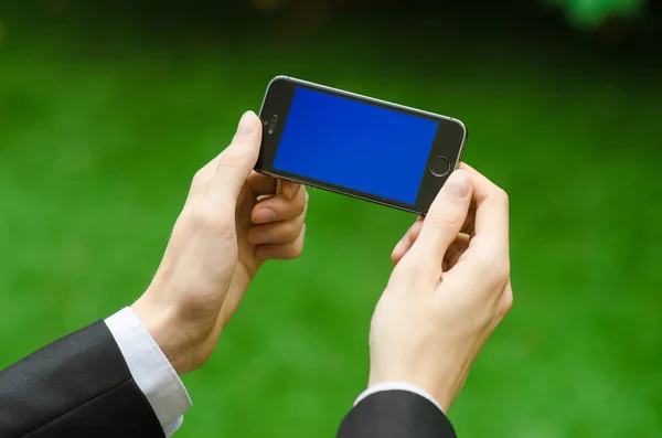 Communication and Business Subject: Hand in a black suit holding a modern phone with blue screen in the background of green grass — Stock Photo, Image