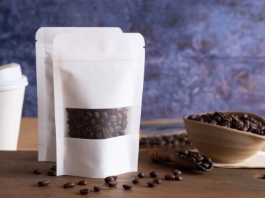 Beyaz şeffaf cam torbasında kavrulmuş kahve çekirdekleri, fermuar ve kase, ahşap arka planda kepçe. Metnin için boşluk.