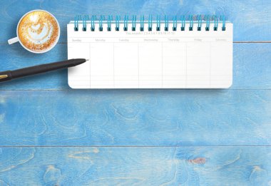 Aylık not defteri ve siyah kalem, modern mavi masabaşı işyerindeki beyaz bardakta kalp şeklinde kahve latte. Üst görünüm, fotokopi alanı ile düz uzanma. İş ve eğitim kavramı