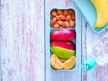 Açık yeşil beslenme çantasında taze meyve, badem, elma, portakal, muz ve açık mavi ahşap arka planda su şişesi. Yemeği götür. Fotokopi alanı, sağlıklı beslenme kavramları