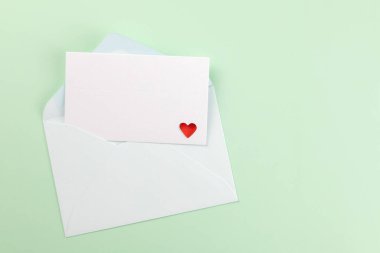 Açık mavi bir zarf, kısmen açık, yeşil arka planda küçük kırmızı bir kalple süslenmiş beyaz bir kart gösteriyor. Aşk mesajlarını, davetiyeleri veya tebrik kartlarını kabul et