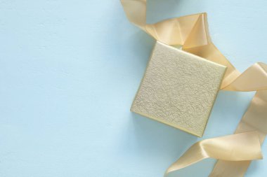 Yumuşak, açık mavi ahşap arka planda saten bir kurdeleyle parıldayan altın bir hediye kutusu. Kutlama için konsept, bayramlar, yıldönümleri ve özel anlar için mükemmel..