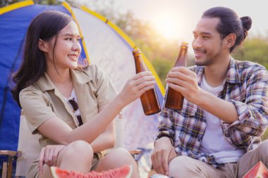 Arkadaşlık Asyalı insanlar kampta içiyorlar, güzel bir kadın elinde bira şişesi ve neşe dolu bir adamla çadırın yanında oturuyorlar, özgürlük ve gün batımında mutlu bir tatil.
