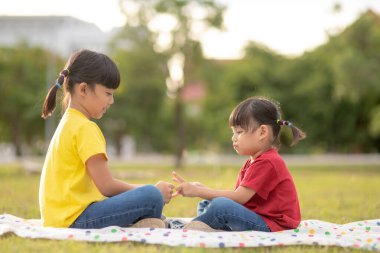 Two little girls sitting on the grass in the park and playing rock paper scissors hand game