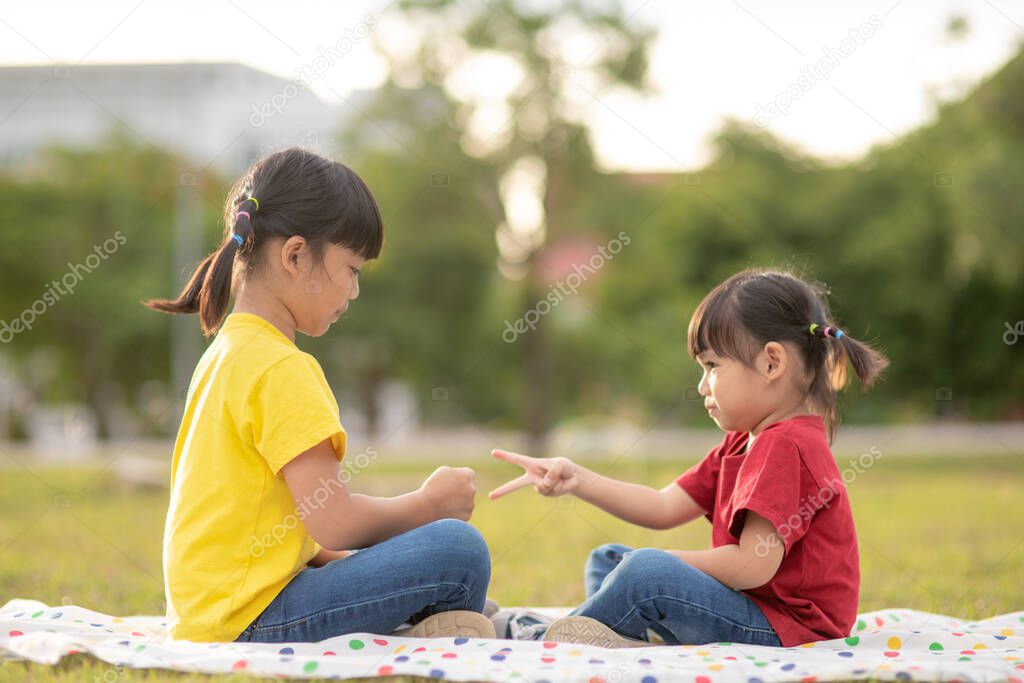 Two little girls sitting on the grass in the park and playing rock paper scissors hand game