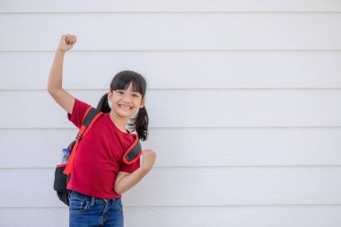 Sırt çantalı neşeli küçük kız atlıyor ve beyaz duvara karşı eğleniyor. Kameraya bakıyorum. Okul konsepti. Okula Dönüş
