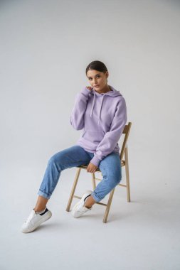 Mor kapüşonlu ve mavi kotlu güzel esmerin portresi gri stüdyo arka planında poz veriyor.