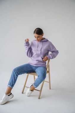 Mor kapüşonlu ve mavi kotlu güzel esmerin portresi gri stüdyo arka planında poz veriyor.