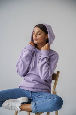 Mor kapüşonlu ve mavi kotlu güzel esmerin portresi gri stüdyo arka planında poz veriyor.