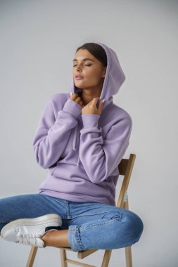 Mor kapüşonlu ve mavi kotlu güzel esmerin portresi gri stüdyo arka planında poz veriyor.
