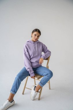 Mor kapüşonlu ve mavi kotlu güzel esmerin portresi gri stüdyo arka planında poz veriyor.