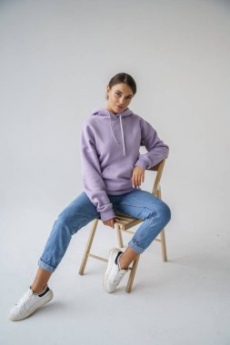 Mor kapüşonlu ve mavi kotlu güzel esmerin portresi gri stüdyo arka planında poz veriyor.