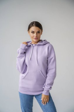 Mor kapüşonlu ve mavi kotlu güzel esmerin portresi gri stüdyo arka planında poz veriyor.