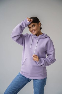 Mor kapüşonlu ve mavi kotlu güzel esmerin portresi gri stüdyo arka planında poz veriyor.