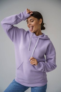 Mor kapüşonlu ve mavi kotlu güzel esmerin portresi gri stüdyo arka planında poz veriyor.