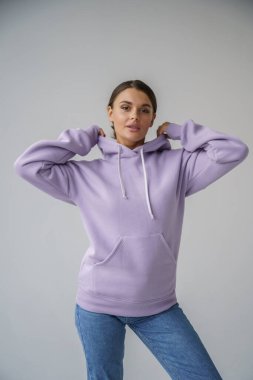 Mor kapüşonlu ve mavi kotlu güzel esmerin portresi gri stüdyo arka planında poz veriyor.