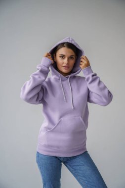 Mor kapüşonlu ve mavi kotlu güzel esmerin portresi gri stüdyo arka planında poz veriyor.