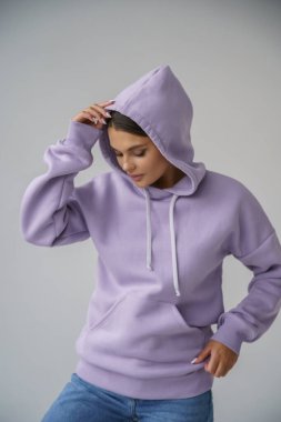 Mor kapüşonlu ve mavi kotlu güzel esmerin portresi gri stüdyo arka planında poz veriyor.