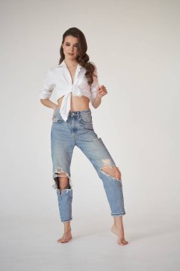 Beyaz gömlekli ve yırtık kot pantolonlu esmer kadın portresi. Beyaz stüdyo arka planında duran genç kadın. 