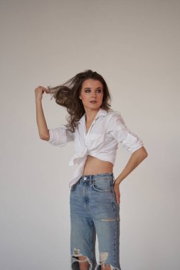 Beyaz gömlekli ve yırtık kot pantolonlu esmer kadın portresi. Beyaz stüdyo arka planında duran genç kadın. 