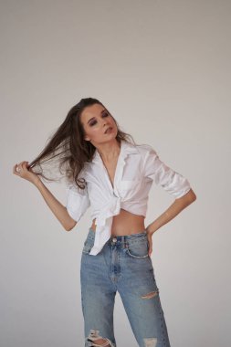 Beyaz gömlekli ve yırtık kot pantolonlu esmer kadın portresi. Beyaz stüdyo arka planında duran genç kadın. 
