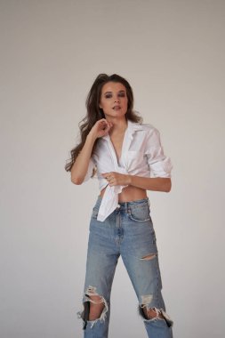 Beyaz gömlekli ve yırtık kot pantolonlu esmer kadın portresi. Beyaz stüdyo arka planında duran genç kadın. 