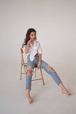 Beyaz gömlekli ve yırtık kot pantolonlu esmer kadın portresi. Tahta sandalyede oturan genç bir kadın, beyaz stüdyo geçmişi. 