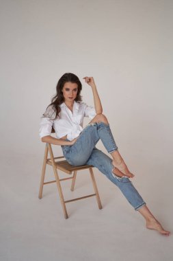 Beyaz gömlekli ve yırtık kot pantolonlu esmer kadın portresi. Tahta sandalyede oturan genç bir kadın, beyaz stüdyo geçmişi. 