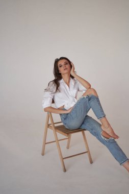 Beyaz gömlekli ve yırtık kot pantolonlu esmer kadın portresi. Tahta sandalyede oturan genç bir kadın, beyaz stüdyo geçmişi. 