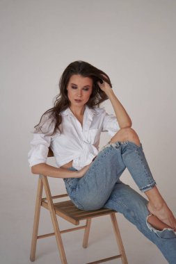 Beyaz gömlekli ve yırtık kot pantolonlu esmer kadın portresi. Tahta sandalyede oturan genç bir kadın, beyaz stüdyo geçmişi. 