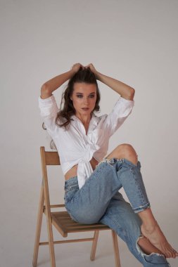 Beyaz gömlekli ve yırtık kot pantolonlu esmer kadın portresi. Tahta sandalyede oturan genç bir kadın, beyaz stüdyo geçmişi. 