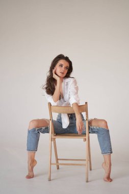 Beyaz gömlekli ve yırtık kot pantolonlu esmer kadın portresi. Tahta sandalyede oturan genç bir kadın, beyaz stüdyo geçmişi. 