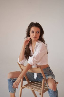 Beyaz gömlekli ve yırtık kot pantolonlu esmer kadın portresi. Tahta sandalyede oturan genç bir kadın, beyaz stüdyo geçmişi. 